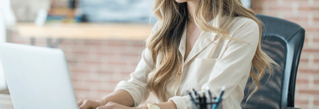 woman sending email on laptop at desk in office with an exposed brick wall behind her