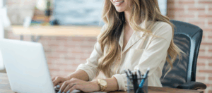 woman sending email on laptop at desk in office with an exposed brick wall behind her